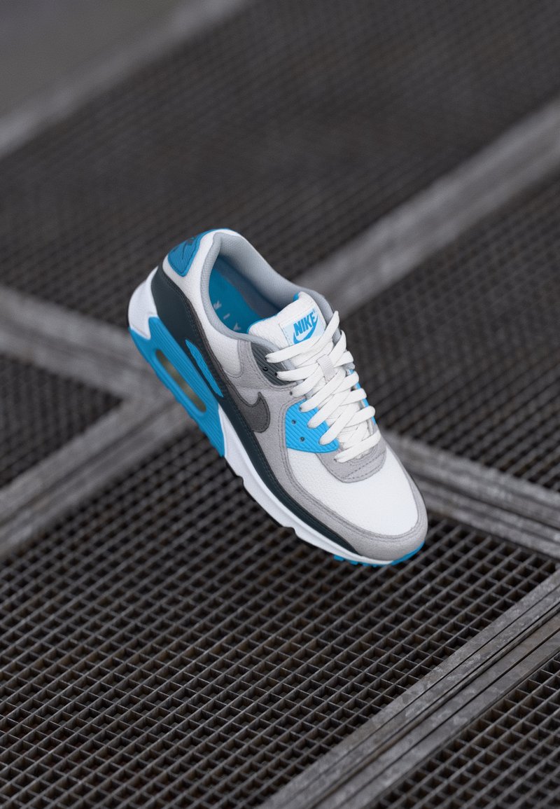 Nike Air Max 90 sneaker featuring a white mesh upper, grey suede accents, light blue highlights, and a visible air cushioning unit.