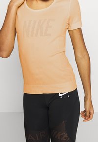 Hedvábně růžový, krátký rukáv, sportovní top s texturovaným povrchem a velkým perforovaným logem "NIKE" na přední straně, doplněný černými legínami se síťovými detaily.