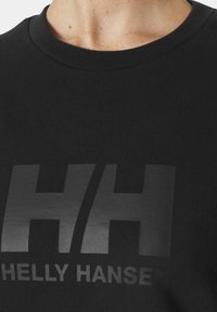 Camiseta de algodón negra con cuello redondo, que presenta un gran logo impreso en negro mate "HH" y el nombre de la marca "Helly Hansen" debajo.