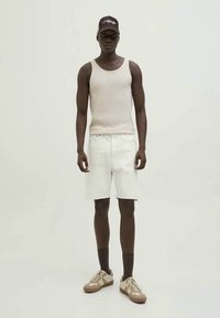 Man in een beige mouwloos ribgebreid T-shirt, witte knielange shorts, donkere sokken, beige sneakers en een donkere pet, staande tegen een effen achtergrond.