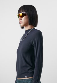 T-shirt à manches longues noir en tissu texturé avec des détails en maille sur les côtés, portant un petit logo près de l'ourlet, associé à des lunettes de soleil réfléchissantes.