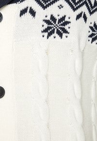 Λευκό πλεκτό καρφίτσωμα με σχέδιο καλωδίων, ναυτικό μπλε λεπτομέρειες και μοτίβα νιφάδας χιονιού. Περιλαμβάνει μαύρα κουμπιά για κλείσιμο.