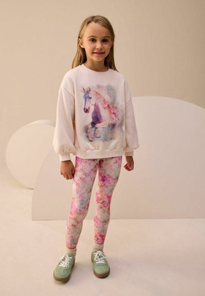 Bleg rosa sweatshirt med et enhjørning grafik, parret med blomster-mønstrede leggings i pastelfarver. Grønne sneakers med hvide snørebånd.
