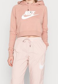 Nike kortärmad hoodie i mjuk rosa med en vit logotyp, tillsammans med ljusrosa byxor med dragsko och vit logotyp på sidan.