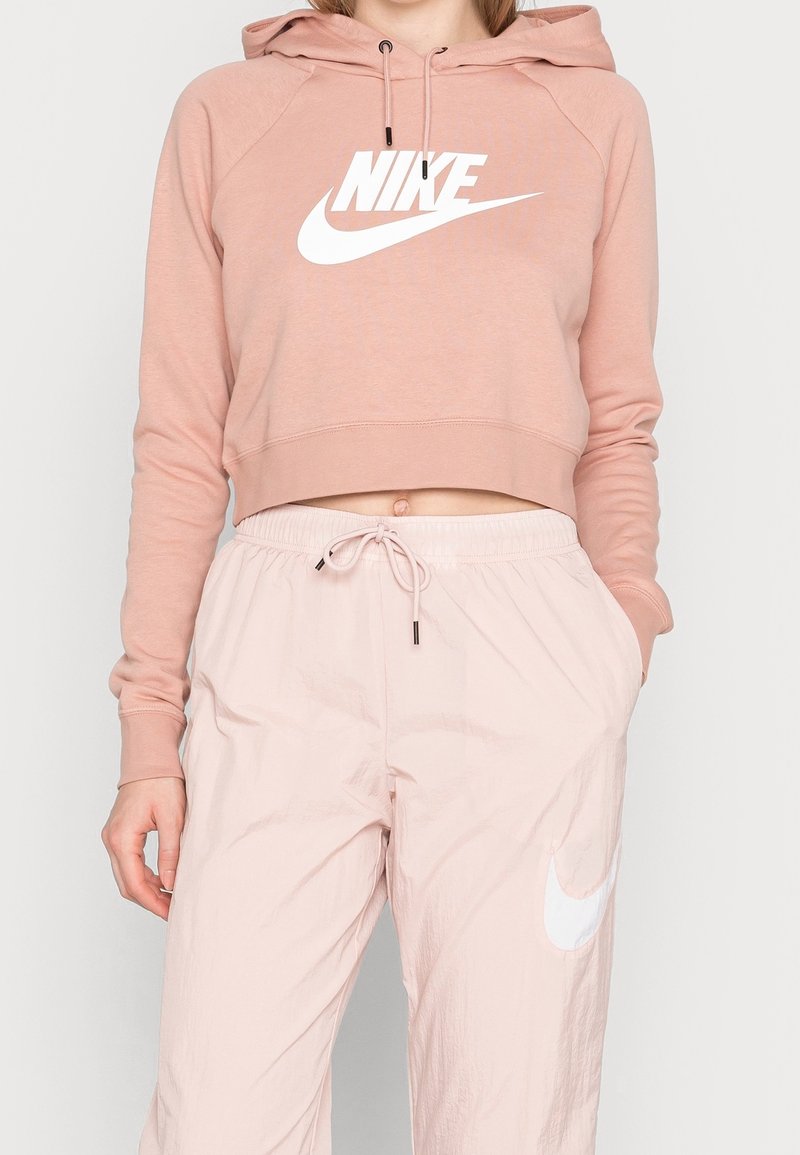 Nike kortärmad hoodie i mjuk rosa med en vit logotyp, tillsammans med ljusrosa byxor med dragsko och vit logotyp på sidan.