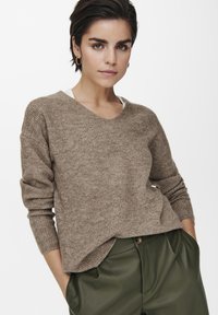 Brauner gerippter Strickpullover mit V-Ausschnitt, langen Ärmeln und lockerem Schnitt, kombiniert mit grünen hochgeschnittenen Hosen.