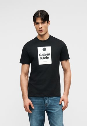 Homme debout portant un t-shirt noir Calvin Klein avec un logo blanc et un jean bleu, sur un fond gris uni.