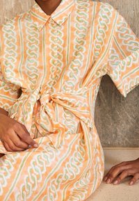Person trägt ein kurzärmliges, pfirsichfarbenes und weiß gemustertes Kleid mit einem vorderen Knoten, ruhend mit einer Hand auf dem Schoß und der anderen auf einer beigen Oberfläche.