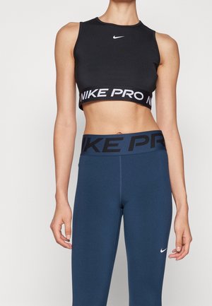 Femme portant un crop top noir Nike Pro et des leggings bleus Nike Pro avec logos visibles sur les vêtements sur un fond uni.