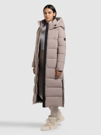 Lange beige donzen jas met capuchon, voorzien van een ritssluiting aan de voorkant, zwarte accenten en ritsen aan de zijkanten. Gecombineerd met beige sportschoenen.