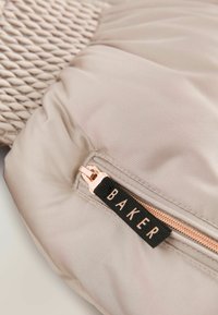 Cojín con tela beige, textura acolchada y una cremallera con tirador de oro rosa. La etiqueta dice "BAKER" en fuente negra sobre un fondo verde.