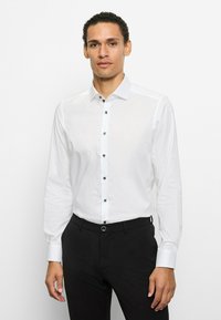 Weißes Langarmshirt mit spitzem Kragen, schwarzen Knöpfen auf der Vorderseite und kontrastierenden schwarzen Akzenten an den Manschetten.