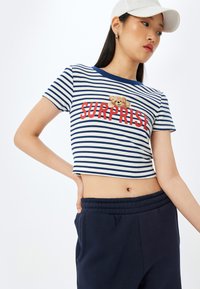 Stribet cropped top i marineblå og hvid med rød "SURPRISE" tekst og et teddybjørne grafik. Bomuldsstof, korte ærmer, ribbet tekstur.