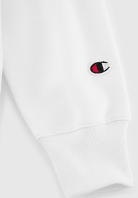 Puño de sudadera blanca en textura acanalada con un pequeño logo bordado en acentos rojos y negros. Tela suave con costuras limpias.