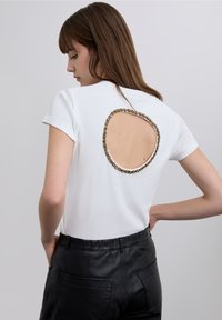 White cotton t-shirt with short sleeves featuring a circular open back adorned with decorative gold-tone beads outlining the cutout.