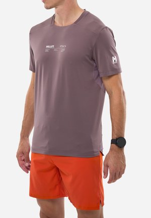 Man wearing purple short-sleeve Millet brand T-shirt and bright orange athletic shorts, with a black smartwatch on left wrist.