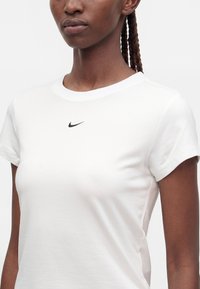 Žena s vrkočmi, ktorá nosí jednoduché biele tričko Nike s malým čiernym logom swoosh uprostred hrude.