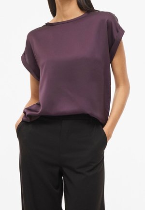 Femme portant un chemisier violet à manches courtes retroussées, rentré dans un pantalon noir taille haute, les mains dans les poches.