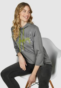 Grijze sweatshirt met een capuchon, voorzien van neon groene koordjes en een grafisch ontwerp in zilver. Gecombineerd met donkergrijze spijkerbroeken.