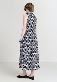 Frau in einem ärmellosen marineblauen Kleid mit weißem geometrischem Muster, schwarzen flachen Schuhen, blauer Armbanduhr und dunkler Sonnenbrille, die seitlich steht.