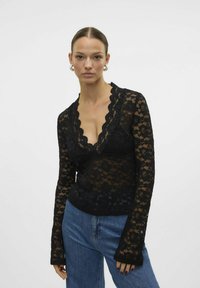 Un haut en dentelle noire à manches longues avec un décolleté en V profond, un motif floral et des bords festonnés, associé à un jean en denim taille haute.