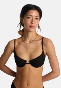 Μαύρο τοπ μπικίνι με ενίσχυση, ρυθμιζόμενους λεπτούς ιμάντες και διακριτικές υφασμάτινες λεπτομέρειες. Το μοντέλο έχει τα μαλλιά του χτενισμένα με ένα αντίθετο φουλάρι.