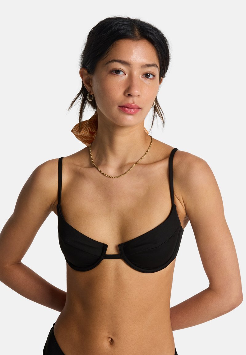 Μαύρο τοπ μπικίνι με ενίσχυση, ρυθμιζόμενους λεπτούς ιμάντες και διακριτικές υφασμάτινες λεπτομέρειες. Το μοντέλο έχει τα μαλλιά του χτενισμένα με ένα αντίθετο φουλάρι.