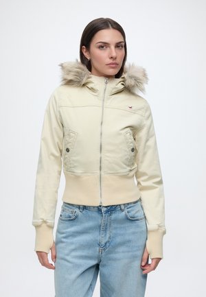 Jeune femme portant une veste beige à capuche avec bordure en fourrure et un jean bleu clair sur un fond blanc uni.