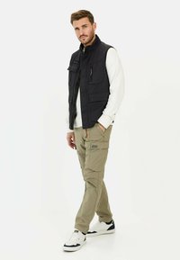 Schwarze ärmellose Weste über einem weißen Pullover, kombiniert mit hellen khakifarbenen Cargohosen und weißen Sneakers. Verfügt über Taschen und einen Reißverschluss vorne.