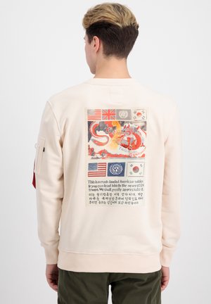 Mand iført cremefarvet sweatshirt med grafik af flag, en drage og tekst, der refererer til en styrtet amerikansk soldat og FN-tropper på ryggen.