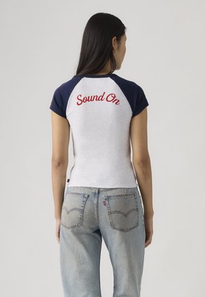 Γυναίκα με μακριά σκούρα μαλλιά που φοράει λευκό και ναυτικό μπλε πουκάμισο με μανίκια ράγκλαν και την επιγραφή "Sound On" με κόκκινα γράμματα στην πλάτη, και ανοιχτόχρωμο μπλε τζιν.