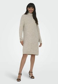 Robe en tricot beige à col roulé, mi-longue avec des manches longues, texture côtelée, fente sur le côté, assortie à des sandales à talons hauts noires à lanières.