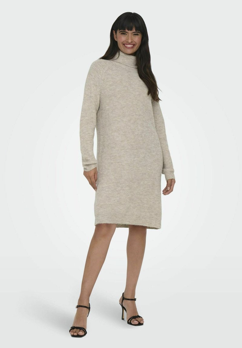 Robe en tricot beige à col roulé, mi-longue avec des manches longues, texture côtelée, fente sur le côté, assortie à des sandales à talons hauts noires à lanières.