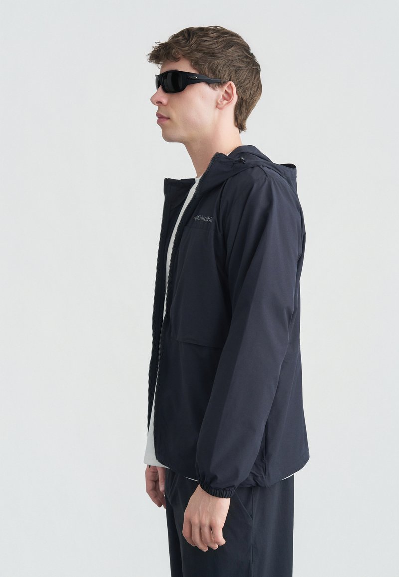 Νεαρός άνδρας που φοράει μαύρα γυαλιά ηλίου, μαύρο φούτερ με κουκούλα και φερμουάρ, λευκό πουκάμισο και μαύρο παντελόνι, στέκεται προφίλ μπροστά σε απλό φωτεινό φόντο.