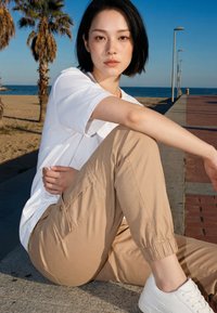 Junge Frau in weißem T-Shirt und beiger Hose sitzt auf einem Gehweg nahe Strandpalmen und Meer unter klarem blauem Himmel.