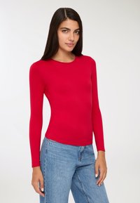 Czerwona bluzka z długim rękawem, wykonana z gładkiego materiału, z okrągłym dekoltem i dopasowanym krojem, w zestawie z niebieskimi dżinsami.
