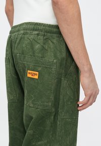 Pantalones cargo verdes hechos de tela texturizada, con una etiqueta de parche y múltiples bolsillos, con cintura elástica y patrón de camuflaje.
