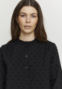 Sort bluse med tekstureret stof og diskrete hævede mønstre, knaplukning og en flæse detalje ved halsen.
