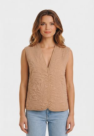 Femme portant un gilet matelassé beige sans manches avec des motifs de feuilles, assorti à un jean bleu clair, debout devant un fond blanc uni.