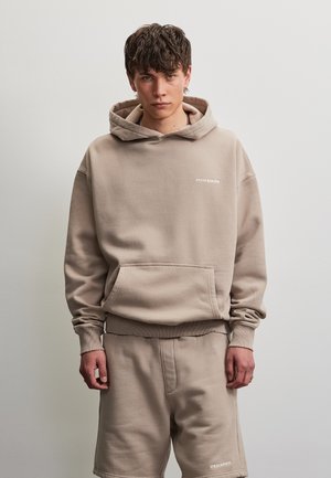 Jeune homme aux cheveux foncés portant un sweat à capuche beige et un short assorti avec le logo "PEGADOR", debout devant un fond clair uni.