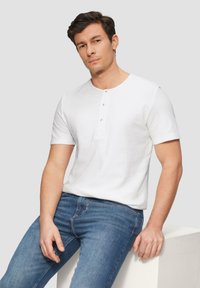 Witte korte mouwen henley-shirt met een placket met drie knopen, gestructureerde stof. Model draagt blauwe slim-fit spijkerbroek. Neutrale achtergrond.