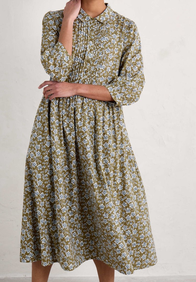 Femme portant une robe midi vert olive à motif floral blanc, manches longues et devant plissé boutonné, debout face à un mur clair uni.