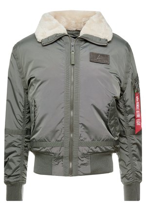 Giacca bomber verde con un morbido colletto in pelliccia sintetica beige, chiusura a zip, polsini elastici e un'etichetta rossa con la scritta "RIMUOVERE PRIMA DEL VOLO."