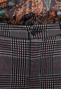 Pantalon à carreaux noir et blanc avec des accents rouges, présentant un tissu texturé, une fermeture à bouton et un haut à motif floral contrasté.