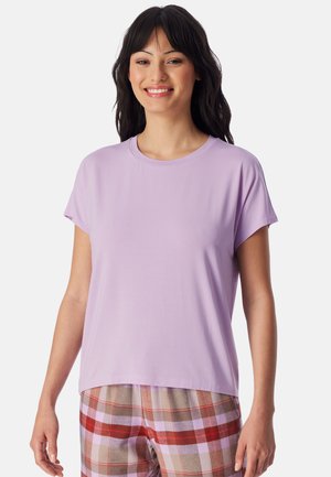 Lächelnde Frau mit langen dunklen Haaren trägt ein helllila kurzärmeliges Shirt und karierte Hose mit Rot-, Beige- und Rosatönen vor weißem Hintergrund.