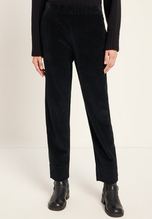 Person wearing black ribbed corduroy pants with cuffed hems and black ankle boots standing against a light background.