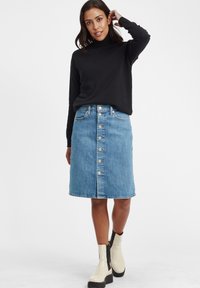 Jeansrock mit einer vorderen Knopfleiste, knielang, in heller blauer Waschung, kombiniert mit einem schwarzen strukturierten Pullover und beigen Knöchelschuhen.
