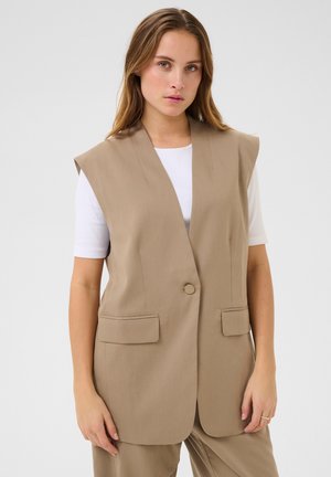 Gilet beige senza maniche con revers a scapestramento, chiusura con un unico bottone, tasche laterali e una texture liscia. Indossato sopra una camicia bianca.