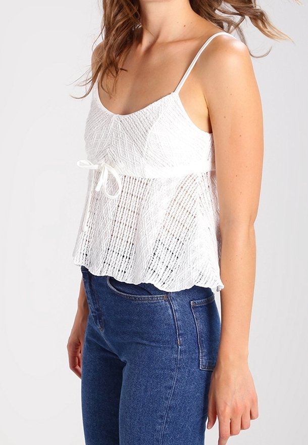 Femme portant un top en dentelle blanche sans manches avec un détail nœud papillon, associé à un jean en denim bleu taille haute, vue de profil.