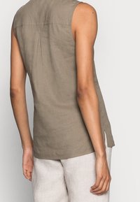 Top senza maniche in lino color marrone chiaro con dettaglio plissettato sulla schiena e fessure sui lati. Abbinato a pantaloni in lino di colore chiaro.